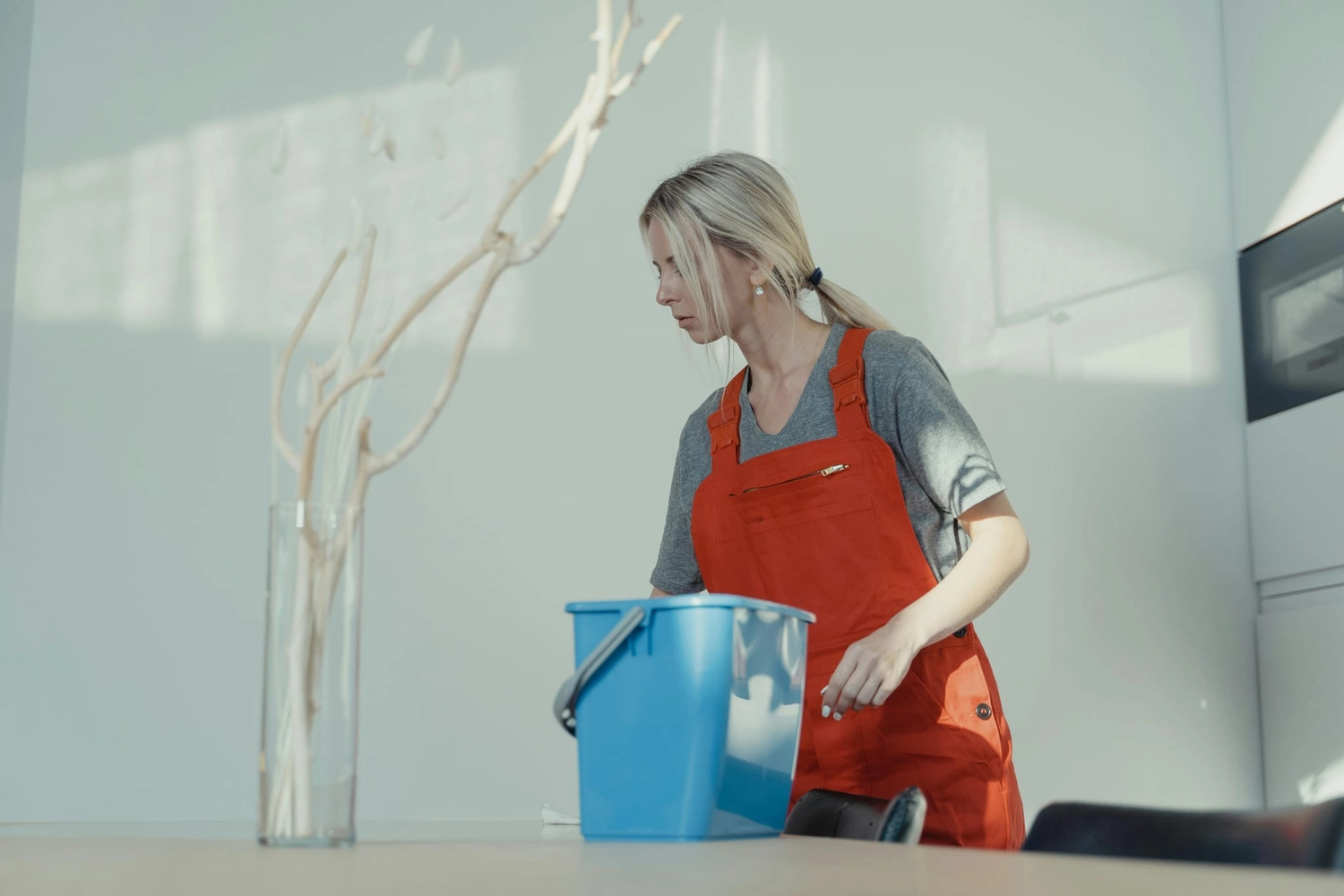 R&O Facilityservice – Eine Frau in einem roten Overall putzt in einem hellen, minimalistisch eingerichteten Raum und hält dabei einen blauen Eimer in der Hand.