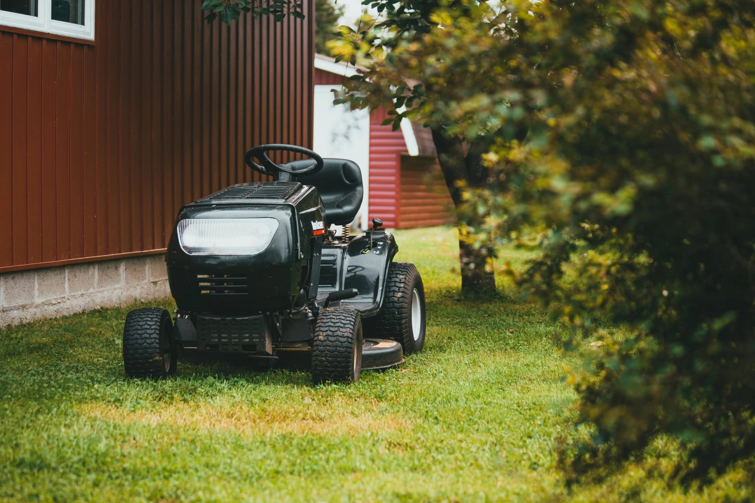 R&O Facilityservice – Ein schwarzer Aufsitzmäher, der auf grünem Rasen neben einer roten Scheune mit Bäumen in der Nähe geparkt ist.