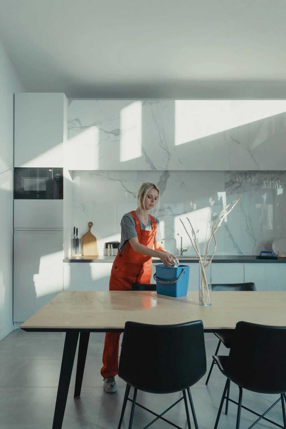 R&O Facilityservice – Eine Frau in einem roten Overall putzt in einem hellen, minimalistisch eingerichteten Raum und hält dabei einen blauen Eimer in der Hand.