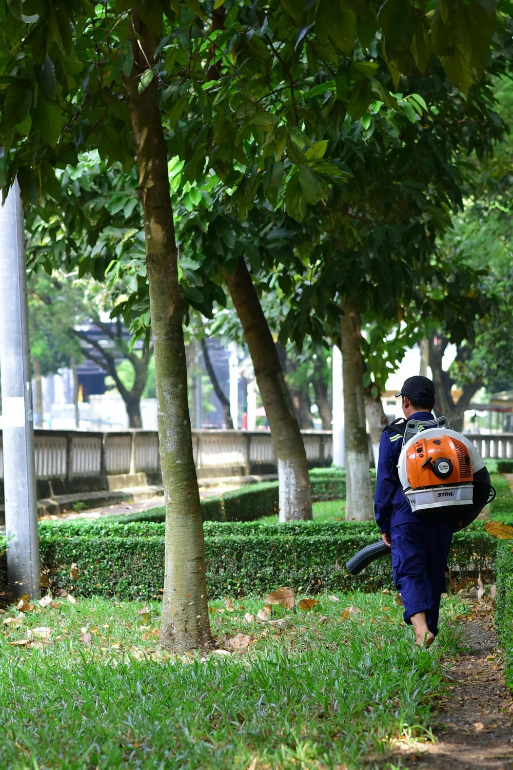 R&O Facilityservice – Eine Person in einem blauen Overall geht mit einem Rucksack-Laubbläser durch einen von Bäumen gesäumten Park.
